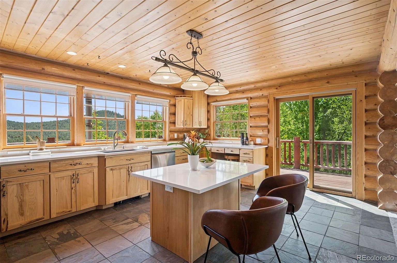 This is a warm and inviting kitchen featuring log walls and ceiling, creating a rustic and cozy atmosphere. The kitchen includes light wood cabinetry, a white countertop island with seating, and stainless steel appliances. Large windows provide ample natural light and views of the surrounding landscape, while a sliding glass door leads to an outdoor deck.