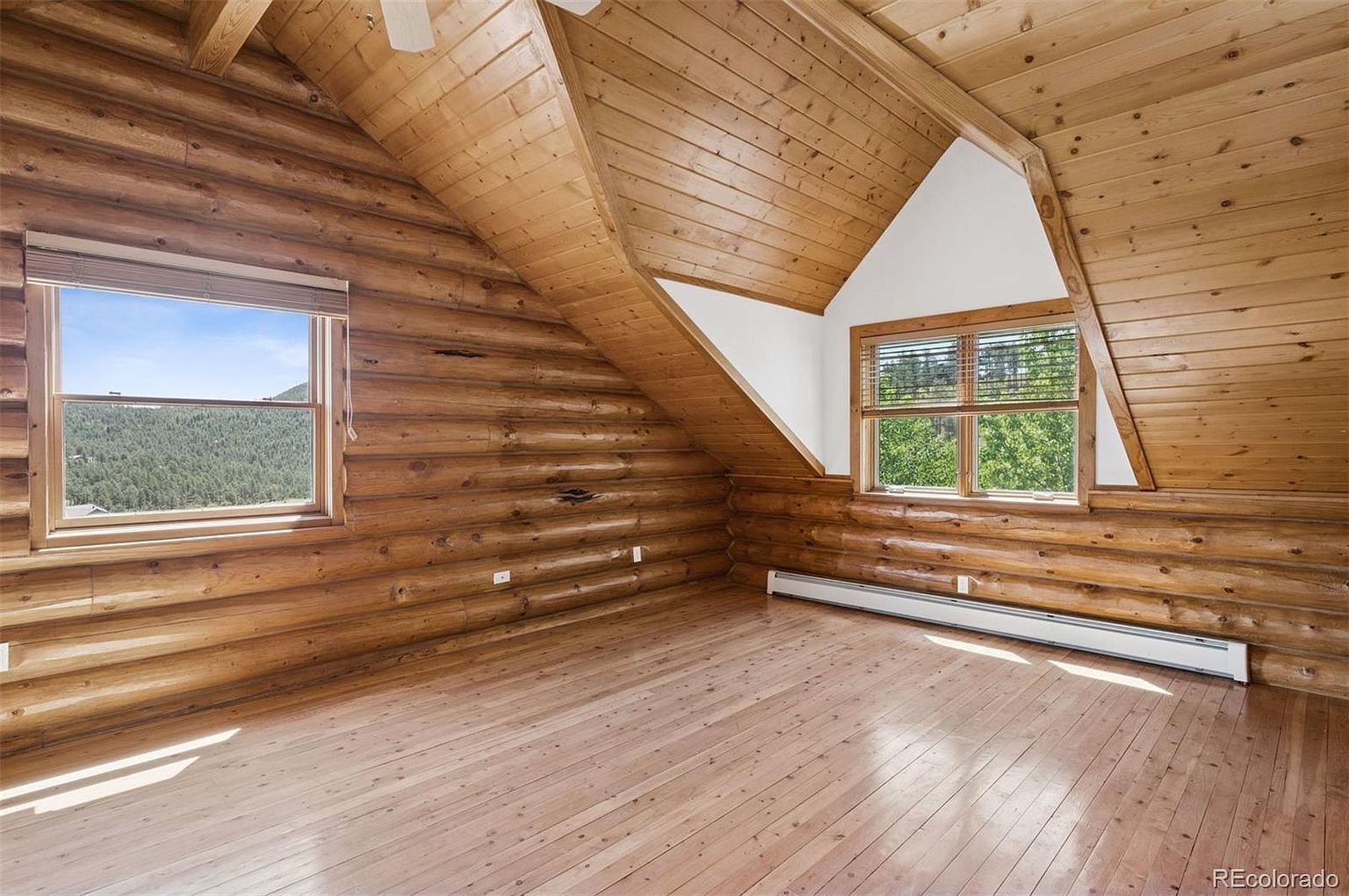 This is an interior shot of a rustic bedroom featuring log walls and ceiling. The room has two windows, one showing a view of trees and the other showing a view of a forest. The hardwood floors are light-colored and the room is spacious and bright, creating a cozy and inviting atmosphere.