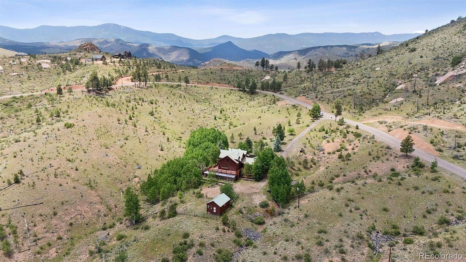 This aerial shot showcases a charming cabin nestled amidst a sprawling landscape of rolling hills and distant mountains. The property features a main house with a deck, a smaller outbuilding, and mature trees providing privacy. A winding road leads to the property, emphasizing its secluded and peaceful setting.