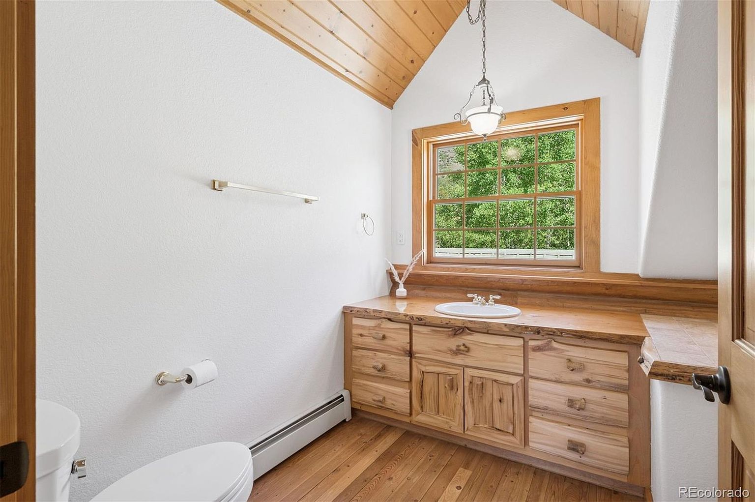 This is a well-lit bathroom featuring a wooden vanity with a built-in sink and multiple drawers and cabinets. A window above the vanity provides natural light and a view of greenery. The walls are white, and the ceiling is wood paneled, creating a warm and inviting atmosphere.