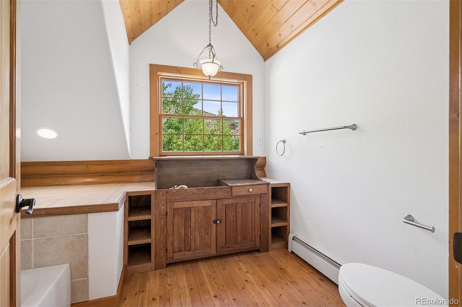 This is a cozy bathroom featuring a wooden vanity with storage, a window providing natural light and a view of greenery, and hardwood floors. The room has a rustic charm with its wooden accents and a sloped ceiling, creating a warm and inviting atmosphere. The toilet is visible in the corner, and a towel rack is mounted on the wall.