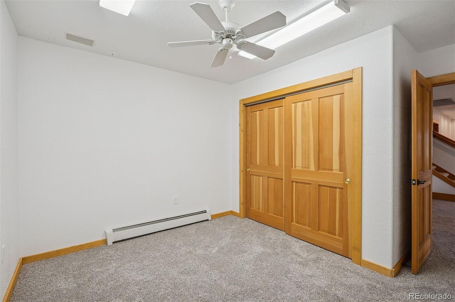 This is a bright and clean bedroom featuring neutral gray carpet, white walls, and a white ceiling fan. A wooden sliding-door closet provides ample storage space, and a doorway leads to another room. The room has a simple and inviting aesthetic, suitable for a guest bedroom.