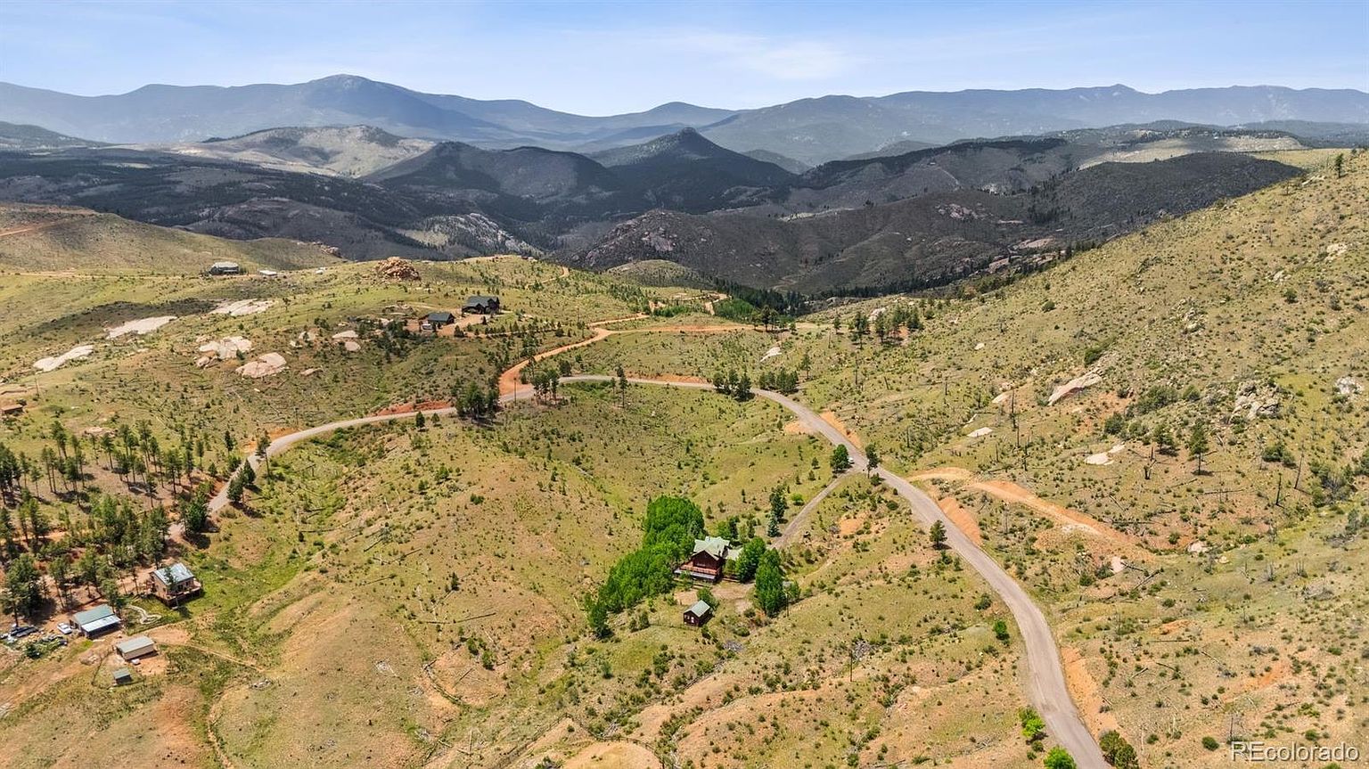This aerial shot showcases a property nestled in a mountainous landscape. A winding road leads to a cluster of buildings surrounded by trees, with rolling hills and distant mountain ranges providing a scenic backdrop. The overall impression is one of seclusion and natural beauty, ideal for a buyer seeking a peaceful retreat.
