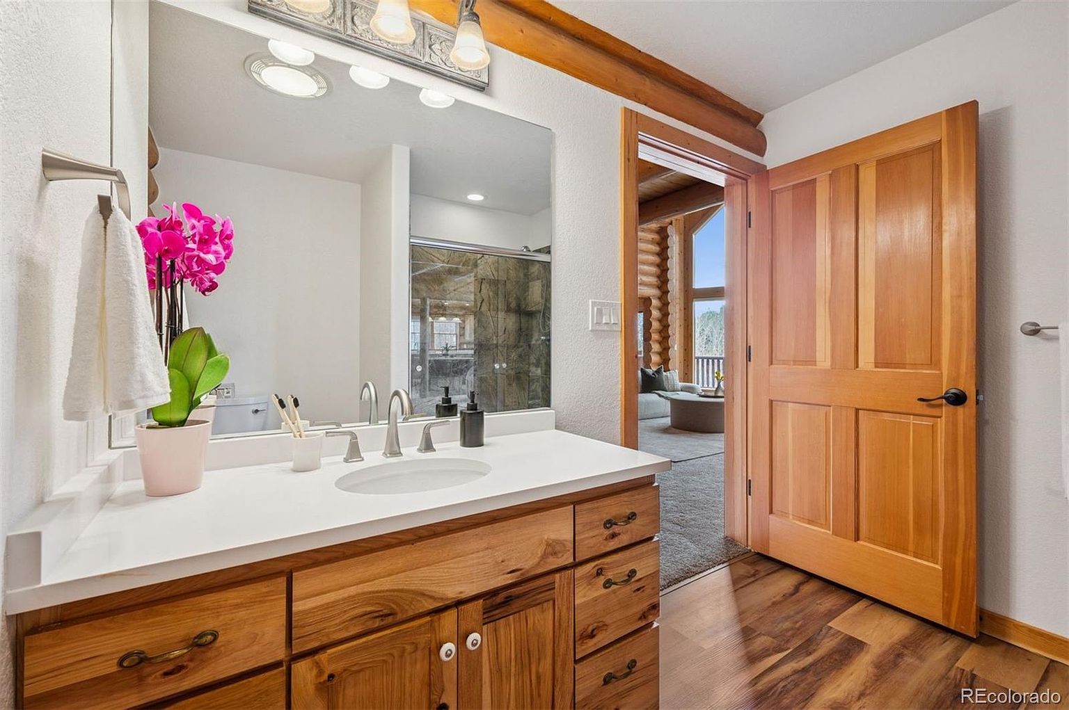 This is a well-lit bathroom featuring a wooden vanity with a white countertop and an oval sink. A large mirror reflects the shower and the adjacent room, creating a sense of spaciousness. The wooden door is slightly ajar, revealing a glimpse of a log cabin-style living area, adding to the rustic charm of the bathroom.
