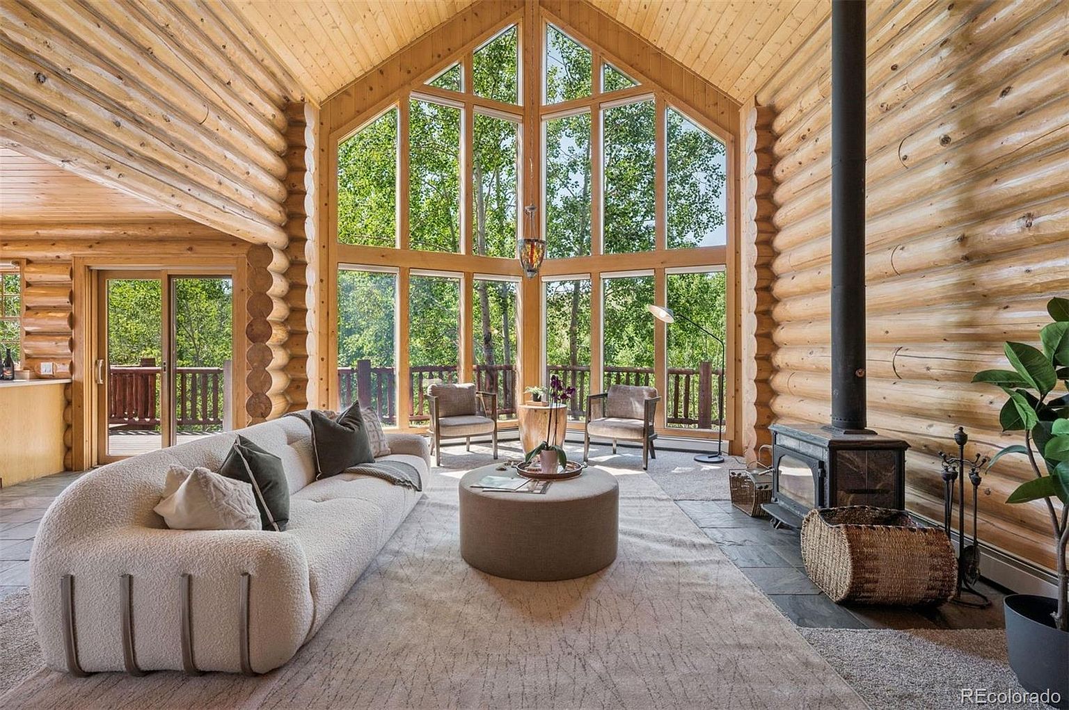 This is an interior shot of a log cabin living room featuring a large window wall with views of the surrounding trees. The room is furnished with a curved sofa, a round ottoman, and two armchairs, creating a cozy and inviting atmosphere. A wood-burning stove adds to the rustic charm of the space.
