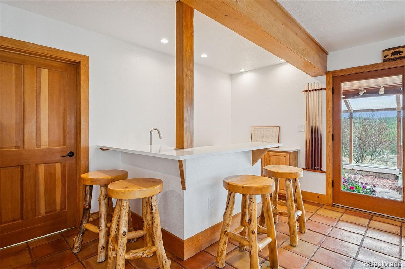 This interior shot showcases a sunroom or conservatory area, featuring a bar with a white countertop and rustic wooden stools. The room has terracotta tile flooring and a wooden door leading to the outside, providing a view of the landscape. The space is well-lit and has a warm, inviting atmosphere.