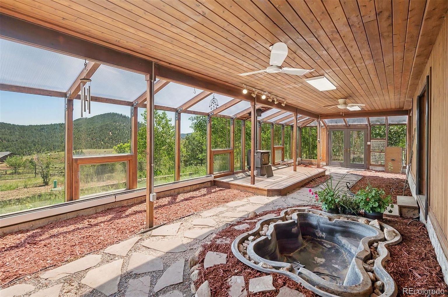 This is an interior shot of a sunroom or conservatory, featuring a wooden ceiling and a glass-enclosed structure that offers views of the surrounding landscape. The room includes a small pond feature, a raised wooden platform with a wood-burning stove, and a stone pathway. The overall impression is a bright, nature-filled space designed for relaxation and enjoyment of the outdoors.
