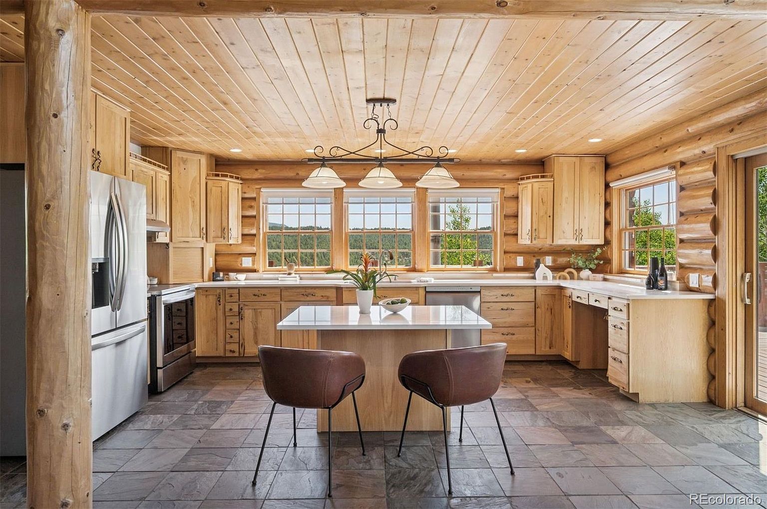 This is a warm and inviting kitchen featuring log cabin construction with wooden walls and ceiling. The kitchen includes light wood cabinetry, stainless steel appliances, a central island with seating, and a large window overlooking a scenic outdoor view. The flooring is tiled, and the overall impression is rustic and cozy.