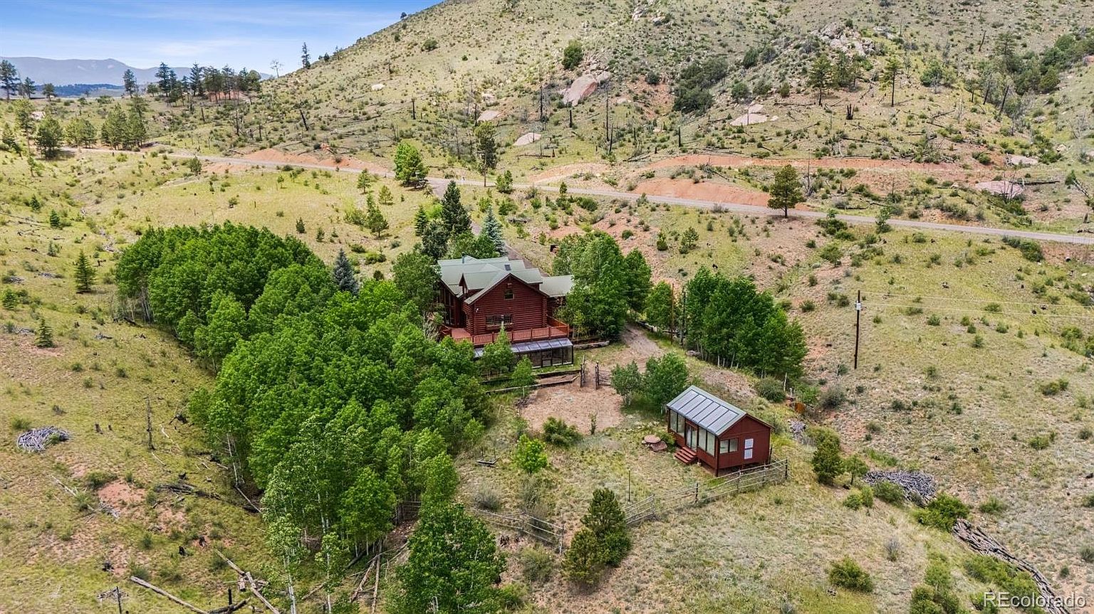 This aerial view showcases a charming log cabin nestled amidst lush greenery and rolling hills. A smaller outbuilding complements the main house, both enclosed by rustic fencing. The property exudes a sense of seclusion and natural beauty, ideal for a serene retreat.