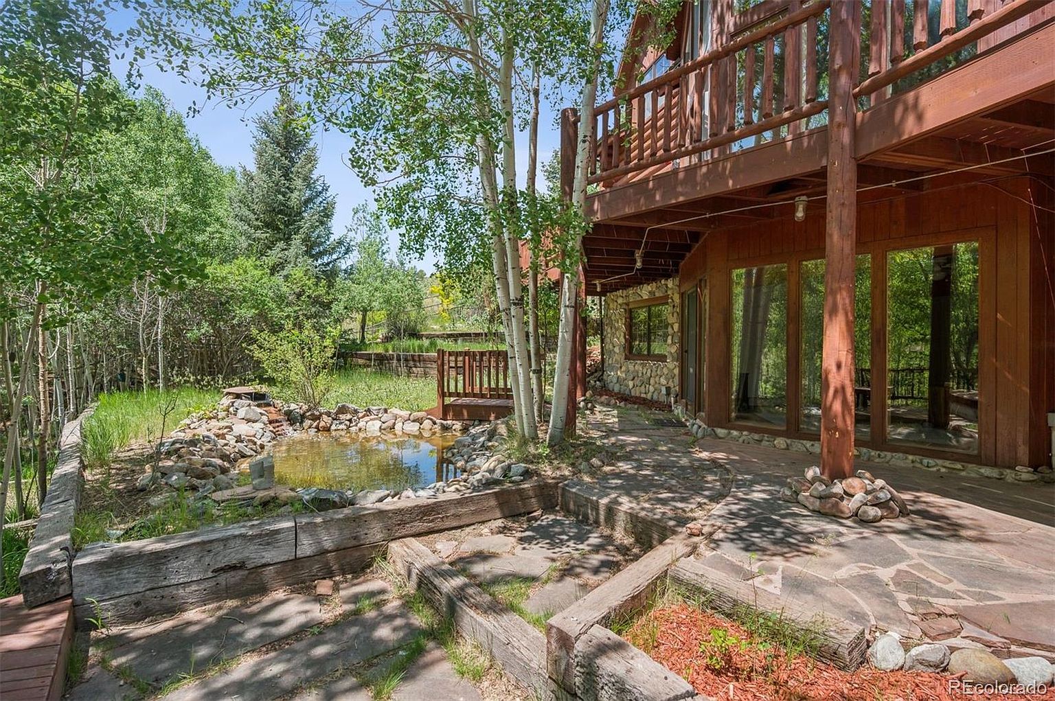 This image showcases a charming outdoor space featuring a multi-level patio and deck area adjacent to a rustic wooden house. A small pond with rock borders adds a natural element, while the wooden deck and stone pathway create an inviting transition between the house and the garden. The overall impression is one of tranquility and connection with nature.