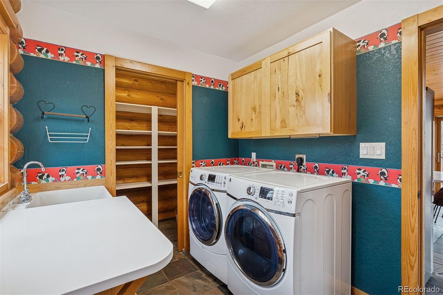 This is a laundry room featuring a front-loading washer and dryer set, light wood cabinets, and a utility sink with a white countertop. The walls are painted a deep teal with a whimsical cow-themed border. A wooden shelving unit is visible through an open doorway, adding to the room's functionality.