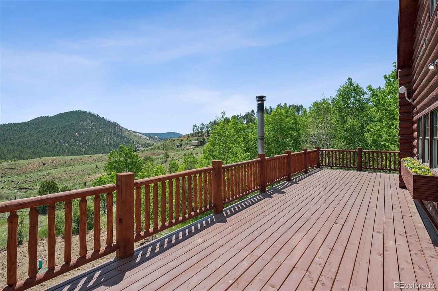 This image showcases a spacious wooden deck with a railing, offering a scenic view of rolling hills and lush greenery under a clear blue sky. The deck appears well-maintained and provides ample space for outdoor seating and entertainment, enhancing the property's appeal for relaxation and enjoying the natural surroundings. A chimney is visible, suggesting a fireplace or stove inside the adjacent building.