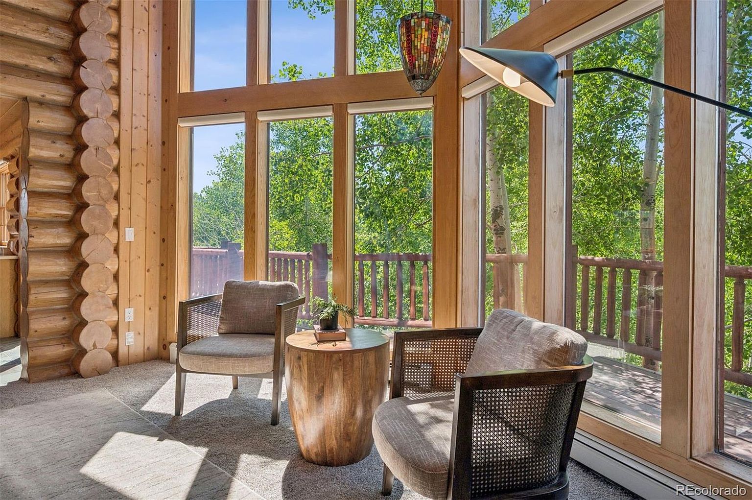 This sunroom features a log wall and a wall of windows that offer a view of the outdoors. Two armchairs and a wooden side table create a cozy seating area. The room is filled with natural light, creating a warm and inviting atmosphere.