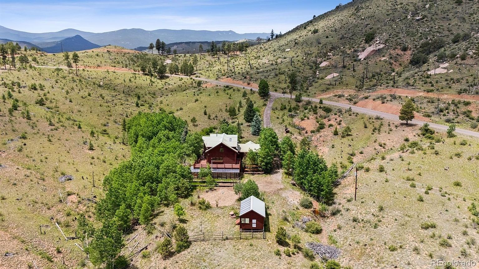 This aerial view showcases a charming cabin nestled amidst a sprawling landscape of rolling hills and scattered trees. The cabin features a wooden exterior, a green roof, and a spacious deck, complemented by a smaller outbuilding nearby. A winding road leads to the property, emphasizing its secluded and peaceful setting.