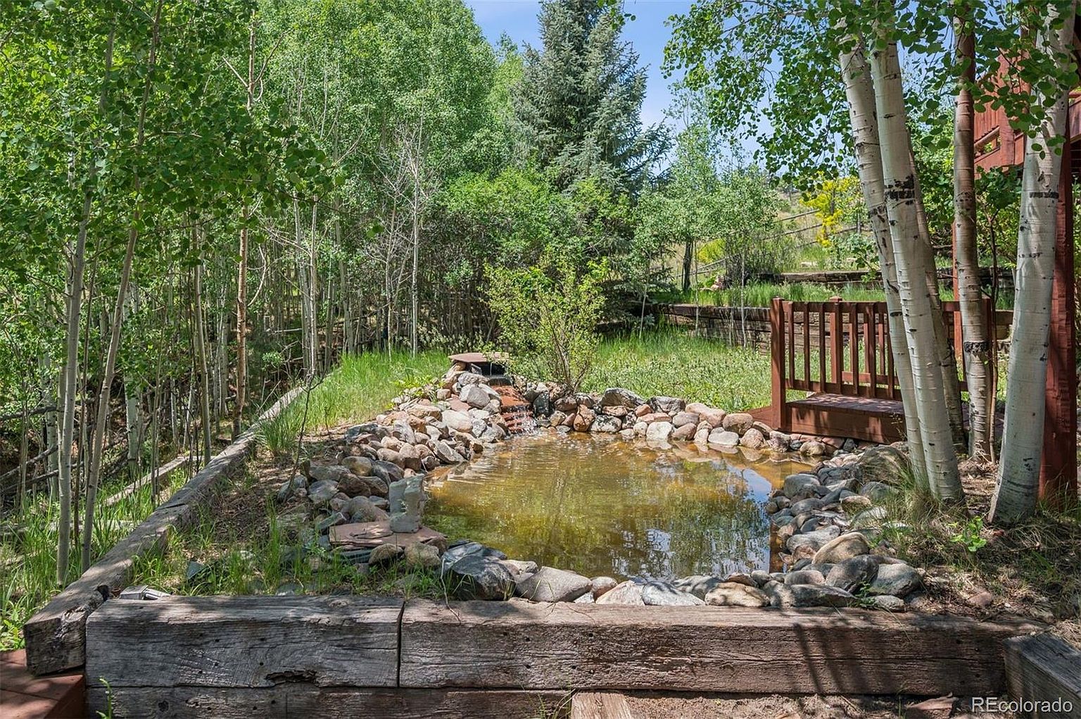 This image showcases a tranquil garden pond surrounded by lush greenery and trees. The pond is lined with rocks and features a small waterfall, creating a serene focal point. A wooden retaining wall and a small deck with a railing add structure and visual interest to the landscape, enhancing the property's appeal.