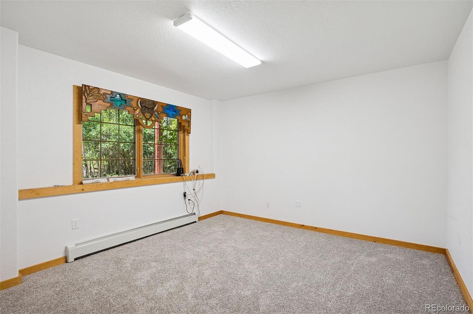 This is a bright, empty bedroom with neutral gray carpet and white walls. A window with a decorative wooden frame provides natural light and a view of the outdoors. The room is clean and ready for personalization, offering a blank canvas for potential buyers.