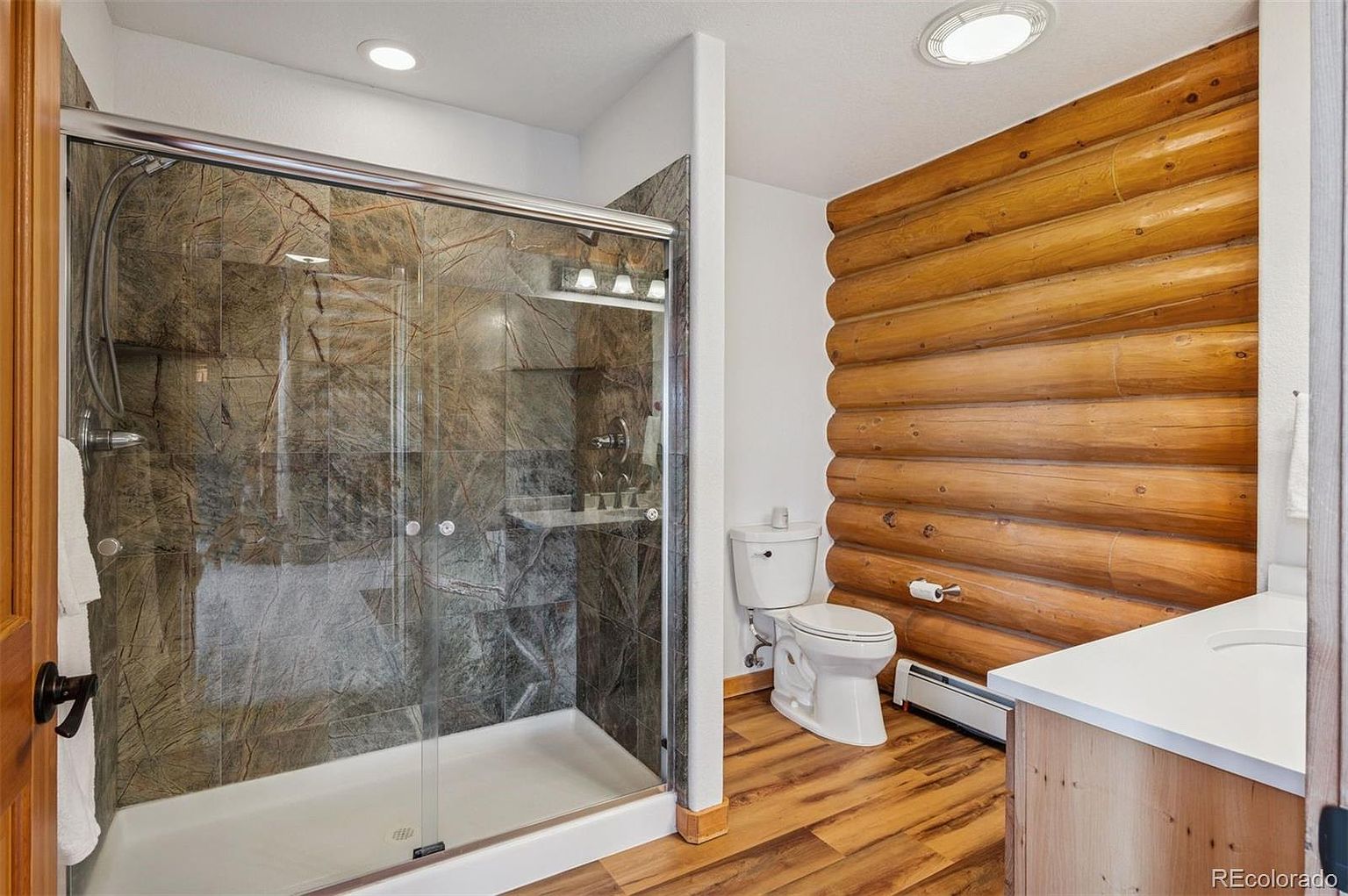 This bathroom features a walk-in shower with glass doors and stone-look tile, a toilet, and a vanity with a white countertop. The wall behind the toilet is constructed of stacked logs, adding a rustic touch. The flooring is wood-look laminate, and the overall style is a blend of modern and rustic.