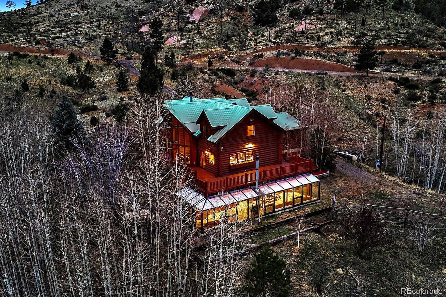 This aerial view showcases a stunning log cabin nestled in a mountainous landscape. The cabin features a green roof, a spacious deck, and a sunroom with glass walls. The surrounding trees add to the property's privacy and natural beauty, making it an ideal retreat.