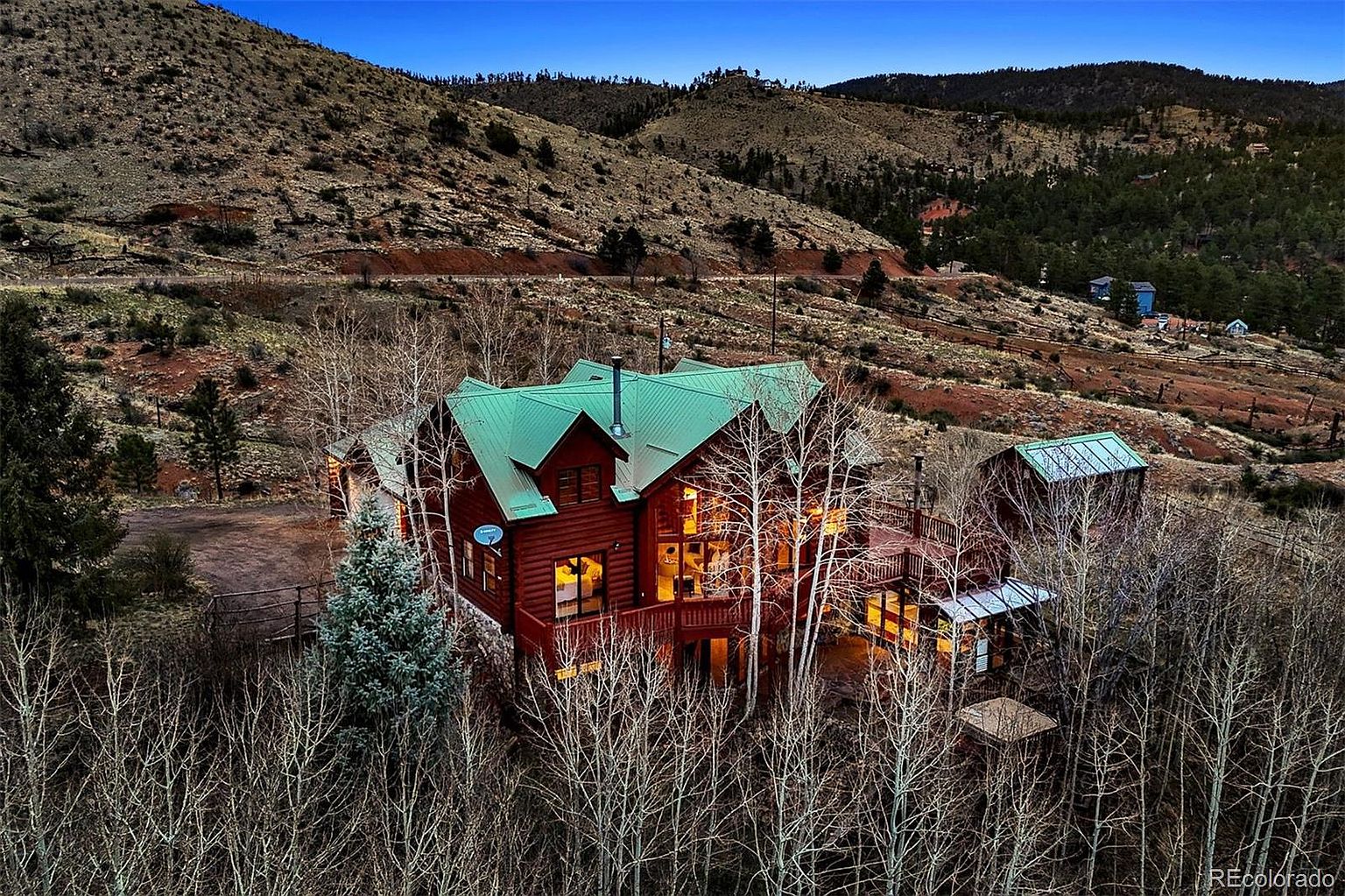 This aerial view showcases a charming log cabin nestled in a mountainous landscape. The cabin features a distinctive green roof and is surrounded by a mix of bare trees and evergreen foliage. The setting provides a sense of seclusion and natural beauty, ideal for a serene retreat.