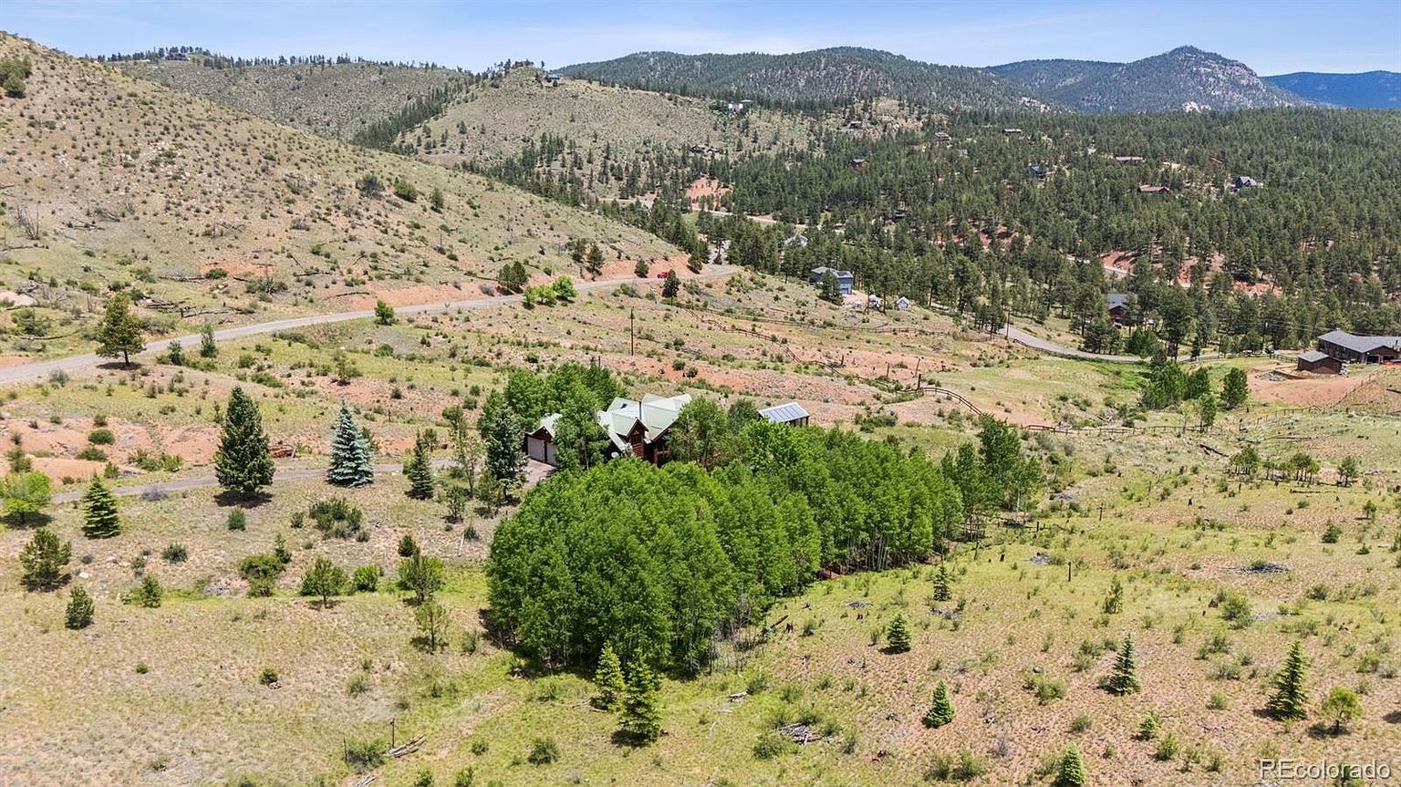 This aerial view showcases a charming home nestled amidst a landscape of rolling hills and scattered trees. The property features a cluster of trees surrounding the house, offering privacy and a sense of seclusion. A winding road leads to the property, highlighting its serene and natural setting.