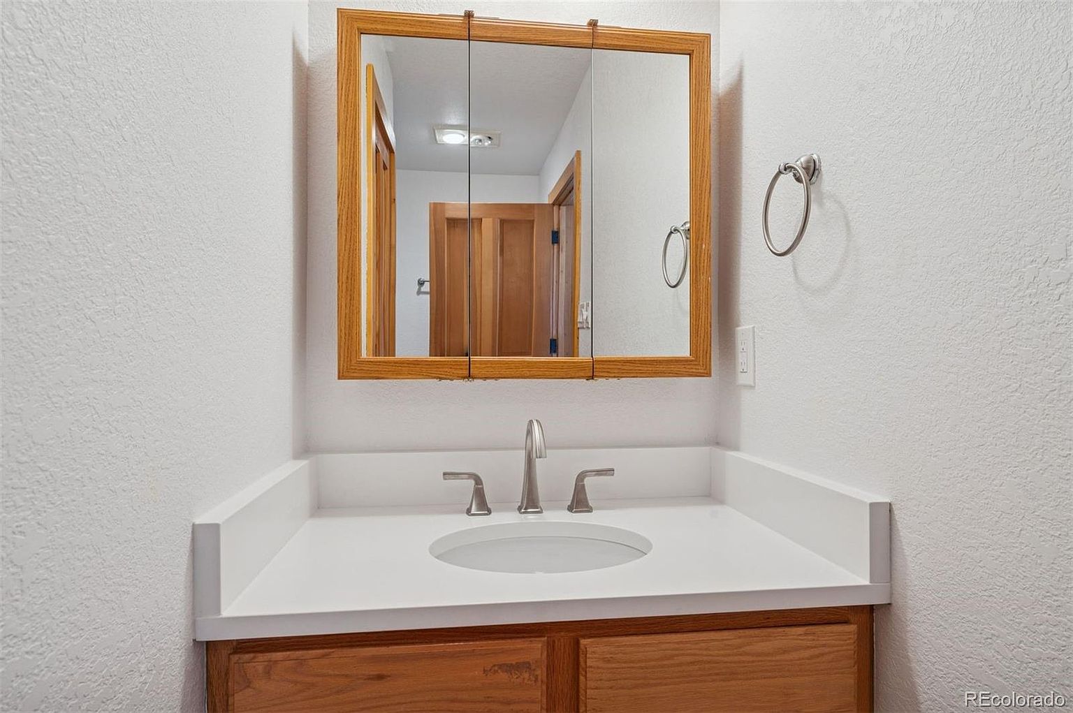 This is a well-lit bathroom featuring a wooden vanity with a white countertop and a circular sink. Above the vanity is a wooden-framed mirror, reflecting a glimpse into another room. A towel ring is mounted on the wall to the right of the mirror, and the walls are painted in a neutral tone, creating a clean and inviting space.