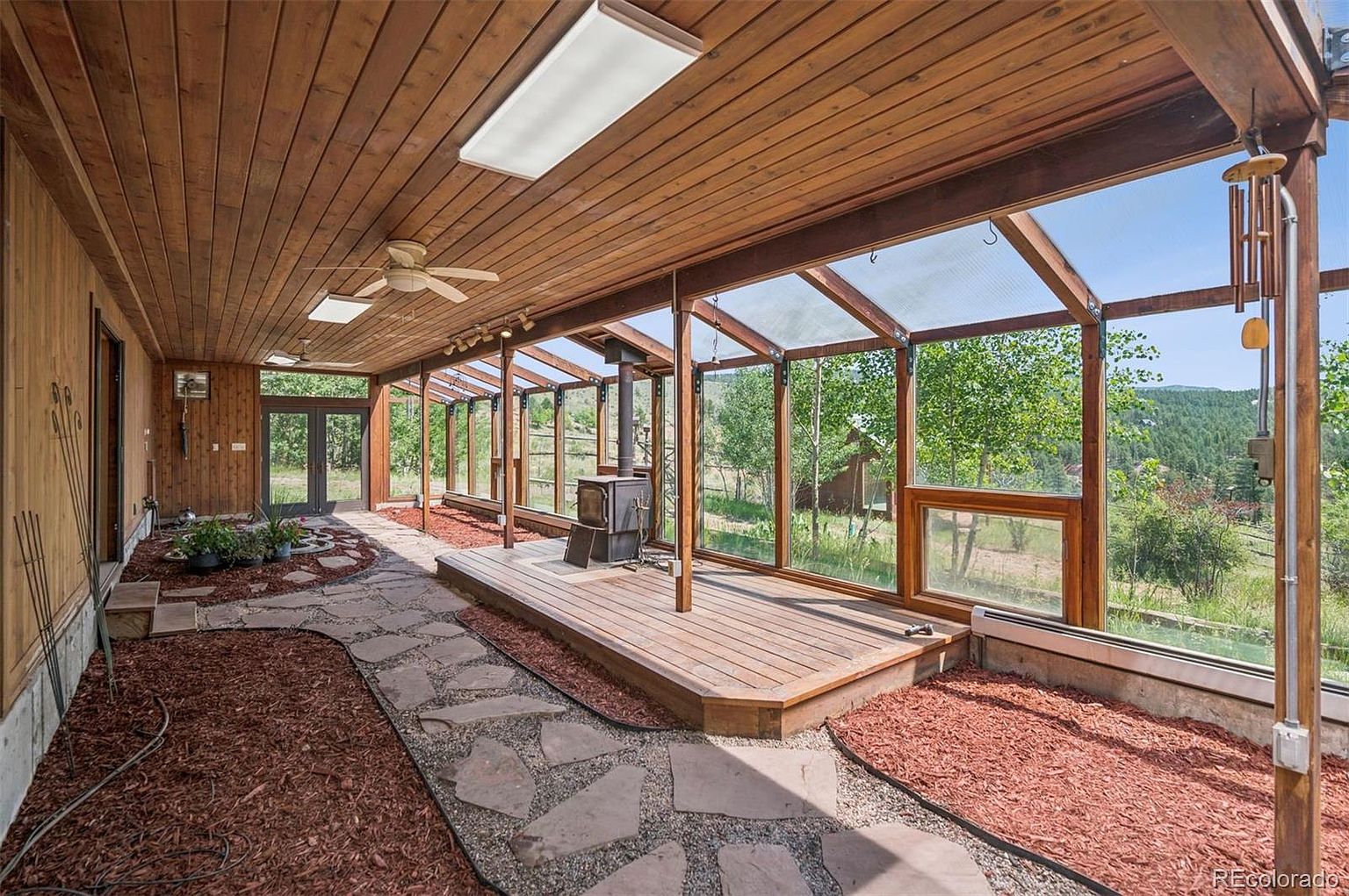This sunroom features a wood-paneled ceiling and a glass-enclosed structure that allows for ample natural light. A wood-burning stove sits on a raised wooden platform, and a stone pathway meanders through the room, adding a rustic touch. The space offers views of the surrounding landscape, creating a serene and inviting atmosphere.