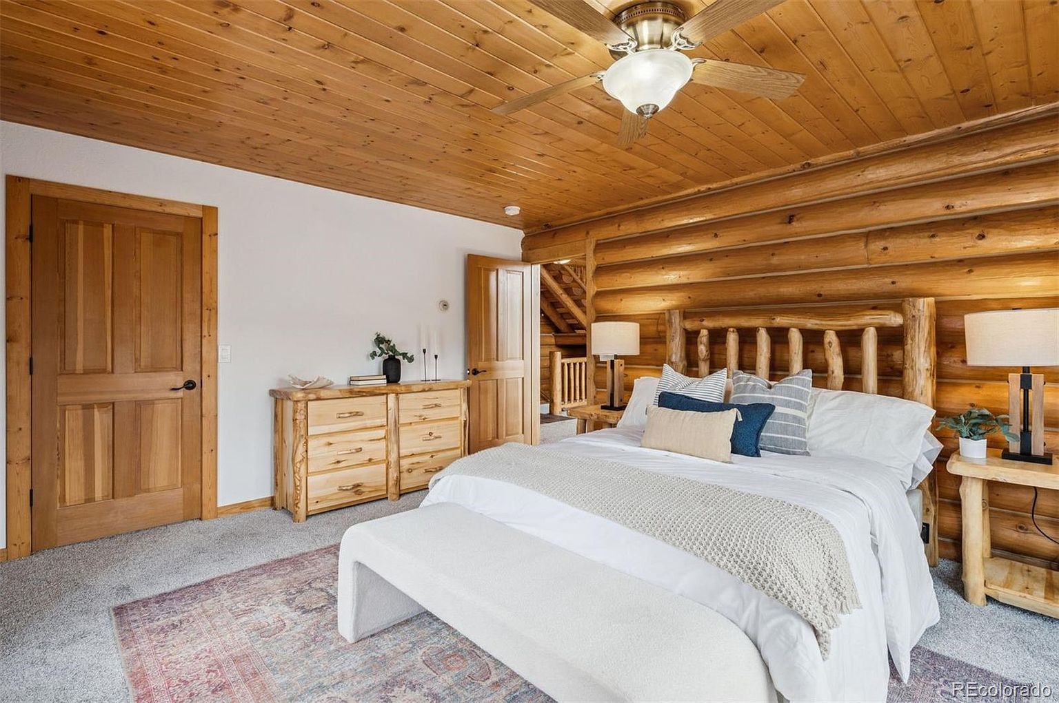 This is a cozy primary bedroom featuring log walls and a wood-paneled ceiling, creating a rustic cabin feel. The room is furnished with a log-style bed frame, a white upholstered bench, and wooden side tables and dresser. A ceiling fan with a light fixture adds to the ambiance, and a patterned rug covers the floor.