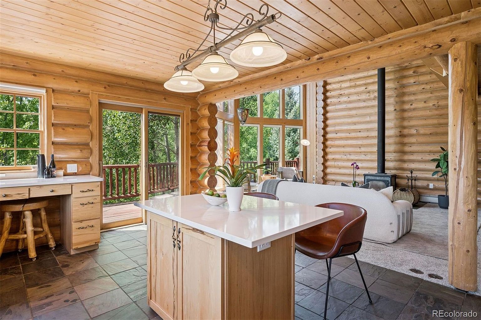 This interior shot showcases a rustic kitchen and living area within a log cabin. The kitchen features a central island with a white countertop, wooden cabinetry, and a unique lighting fixture above. The living area includes a cozy seating arrangement with a fireplace, creating a warm and inviting atmosphere.