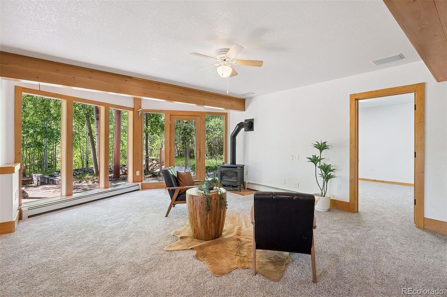 This is an interior shot of a living room featuring a wood-burning stove, large windows with a view of a wooded area, and neutral-toned carpeting. The room is decorated with wooden accents, including beams and door frames, and includes a unique hide rug and modern furniture. The space is bright and airy, creating a cozy and inviting atmosphere.