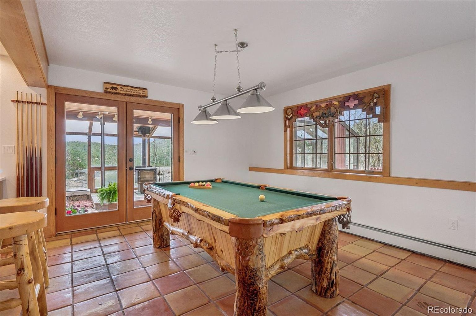 This sunroom features a rustic-style pool table with a green felt top, complemented by unique wooden legs and trim. The room is illuminated by a hanging light fixture above the table and natural light from a window and glass doors leading to an outdoor area. The terracotta tile flooring adds warmth to the space, creating a cozy and inviting atmosphere.