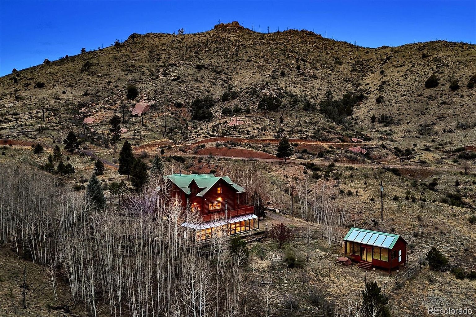 This aerial view showcases a charming log cabin nestled in a mountainous landscape. The property features a main house with a green roof and a smaller outbuilding, surrounded by trees and natural terrain. The setting offers privacy and a connection to nature, making it an appealing retreat.