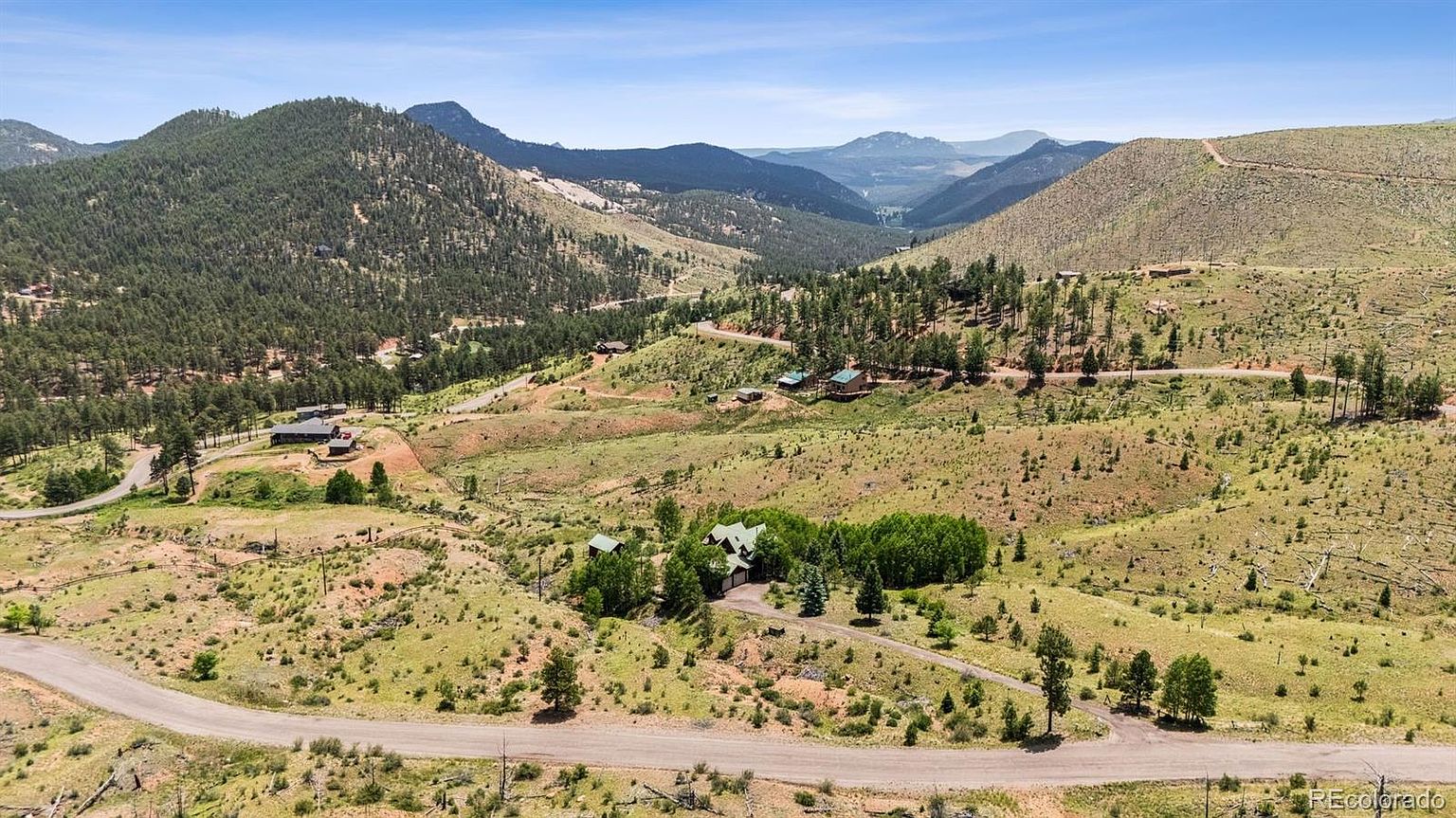 This aerial view showcases a sprawling property nestled in a mountainous landscape. A winding dirt road leads to a house surrounded by lush greenery and mature trees. The setting offers privacy and scenic views, highlighting the property's natural beauty and secluded location.