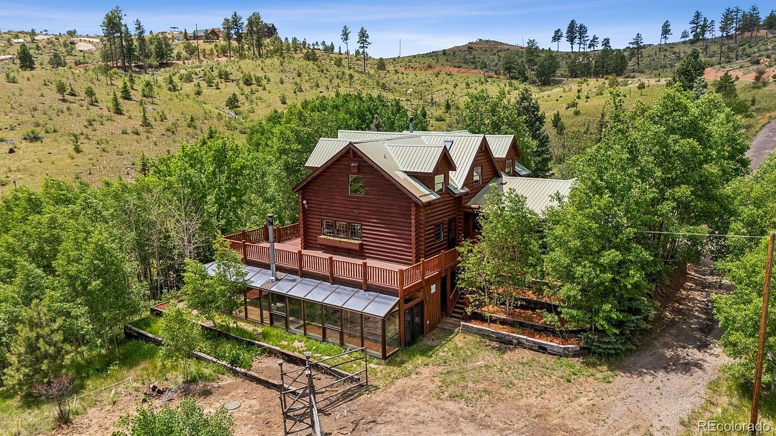 This aerial view showcases a stunning log cabin home nestled amidst lush greenery and rolling hills. The house features a metal roof, a spacious deck, and a sunroom. The surrounding landscape includes mature trees and a well-maintained yard, creating a serene and private setting.
