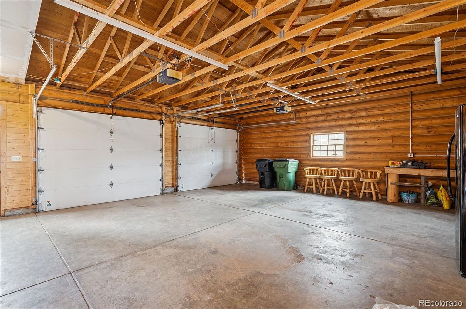 This is an interior shot of a spacious garage featuring two white garage doors and a wooden ceiling with exposed beams. The walls are constructed of wood logs, adding a rustic touch. The concrete floor is expansive and well-lit, providing ample space for vehicles and storage.
