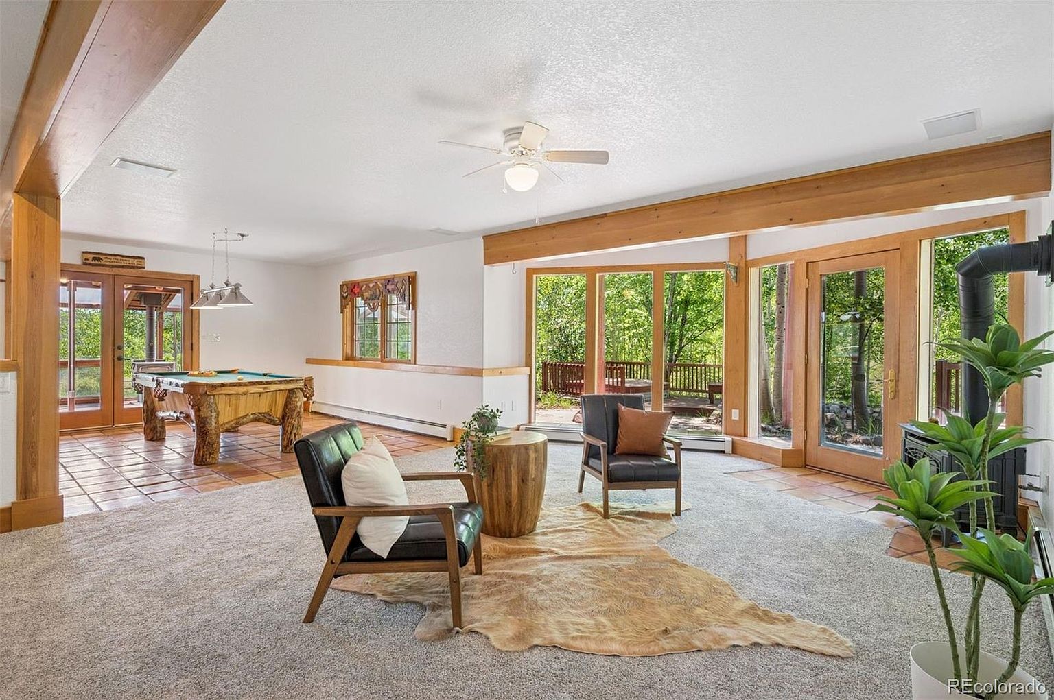 This is an interior shot of a living room featuring a pool table, two armchairs, and a cowhide rug. The room has a rustic-modern style with wooden beams and trim, a mix of tile and carpet flooring, and large windows offering views of the outdoors. A wood-burning stove adds a cozy touch to the space.
