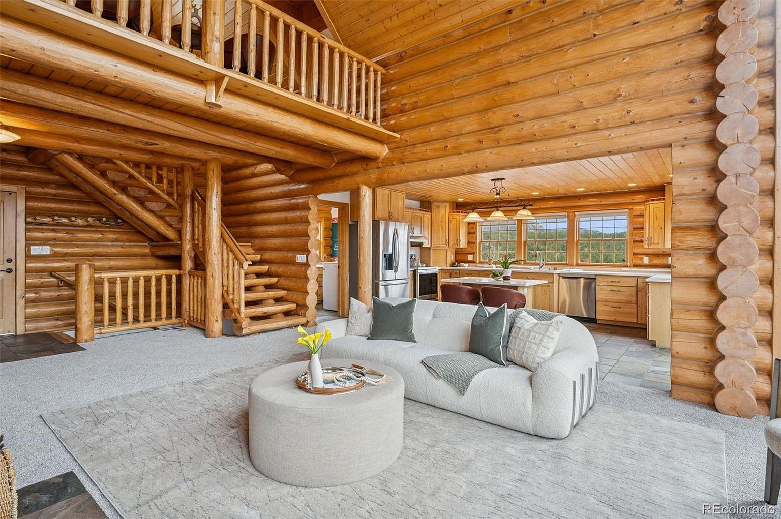 This is an interior shot of a log cabin-style living room that seamlessly transitions into the kitchen. The living room features a large, curved white sofa, a round ottoman, and a light-colored rug. The kitchen has wooden cabinets, stainless steel appliances, and a breakfast bar, creating an open and inviting space with a rustic yet modern feel.