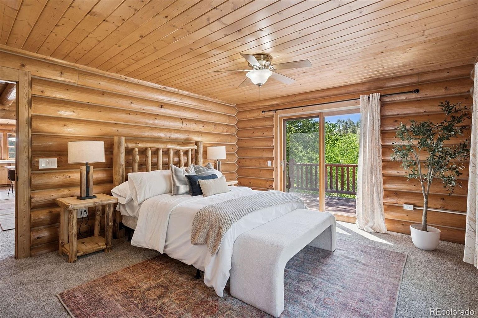 This is a cozy primary bedroom featuring log walls and ceiling, creating a rustic cabin feel. The room is furnished with a bed, bedside tables with lamps, and a bench at the foot of the bed. A sliding glass door leads to a balcony, bringing in natural light and offering a view of the outdoors.