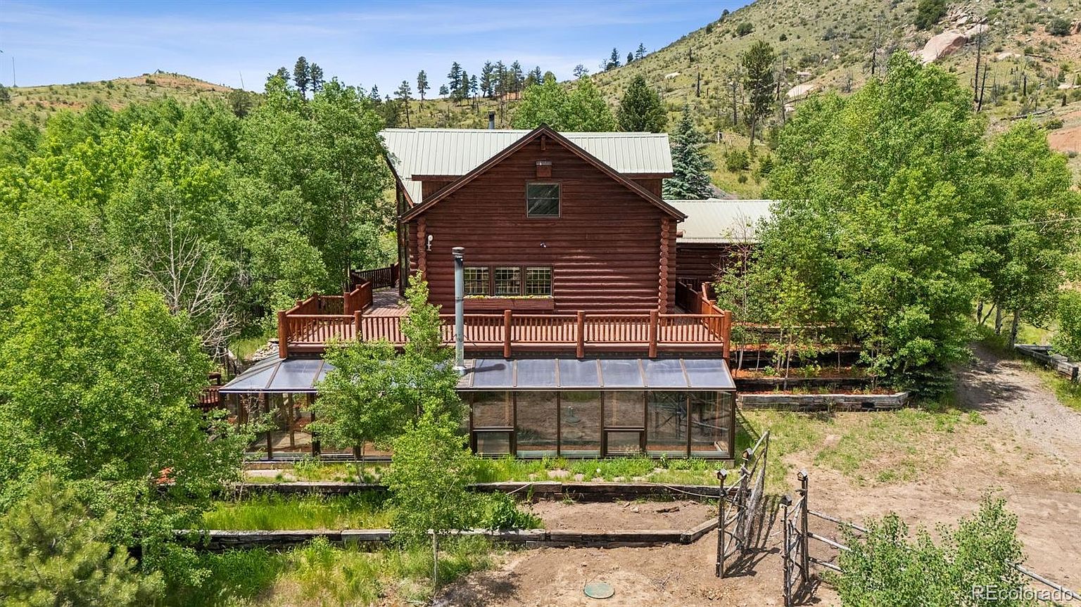 This image showcases the rear exterior of a log cabin home, featuring a spacious wooden deck and an attached greenhouse. The cabin is surrounded by lush greenery and trees, with a mountain visible in the background. The overall impression is one of rustic charm and tranquility, ideal for nature lovers.