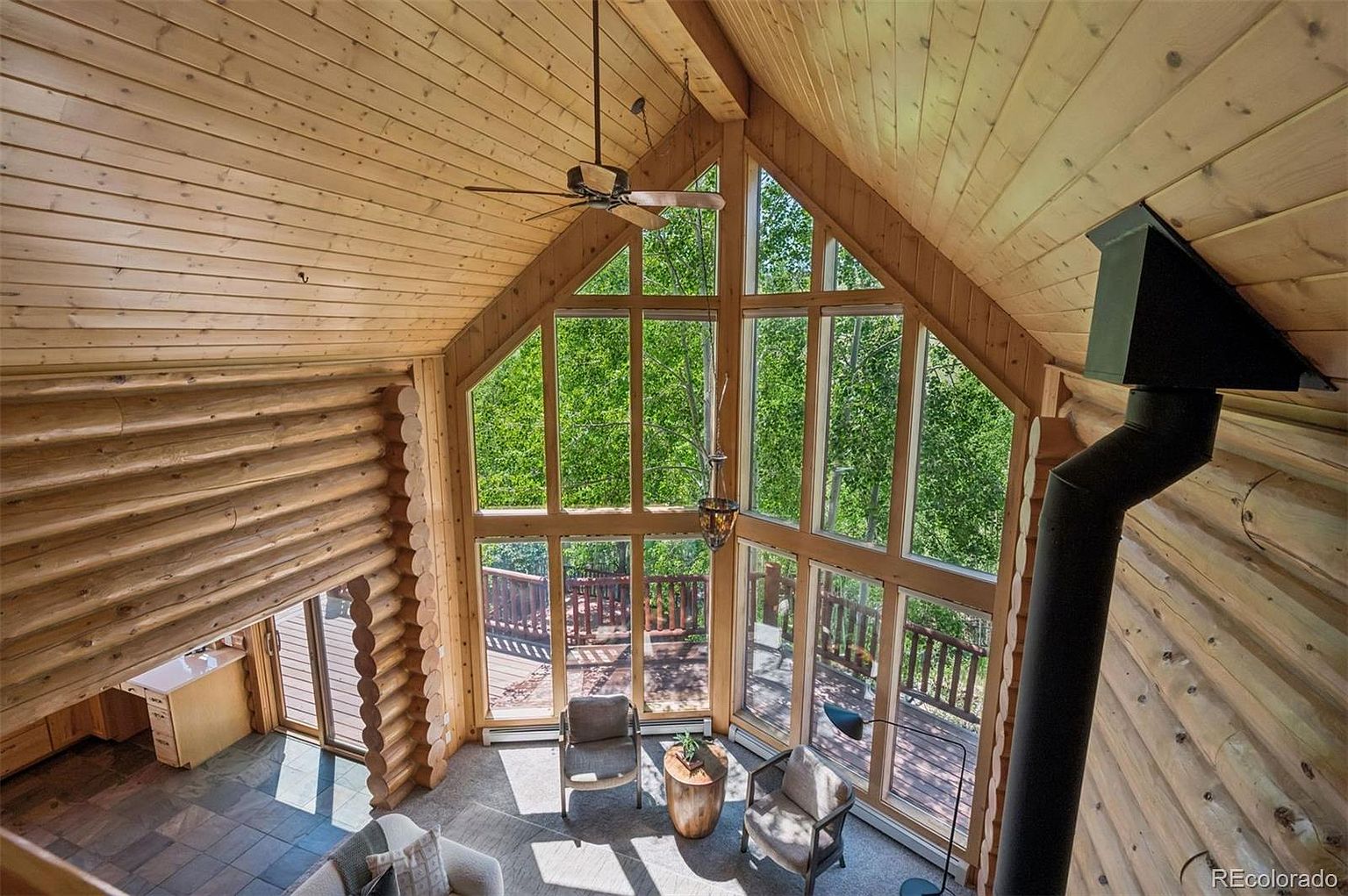 This is an interior shot of a log cabin living room, showcasing a soaring A-frame ceiling and a wall of windows that flood the space with natural light and offer views of the surrounding trees. The room features a cozy seating area with chairs and a small table, complemented by a wood-burning stove with a prominent black chimney, creating a warm and inviting atmosphere.