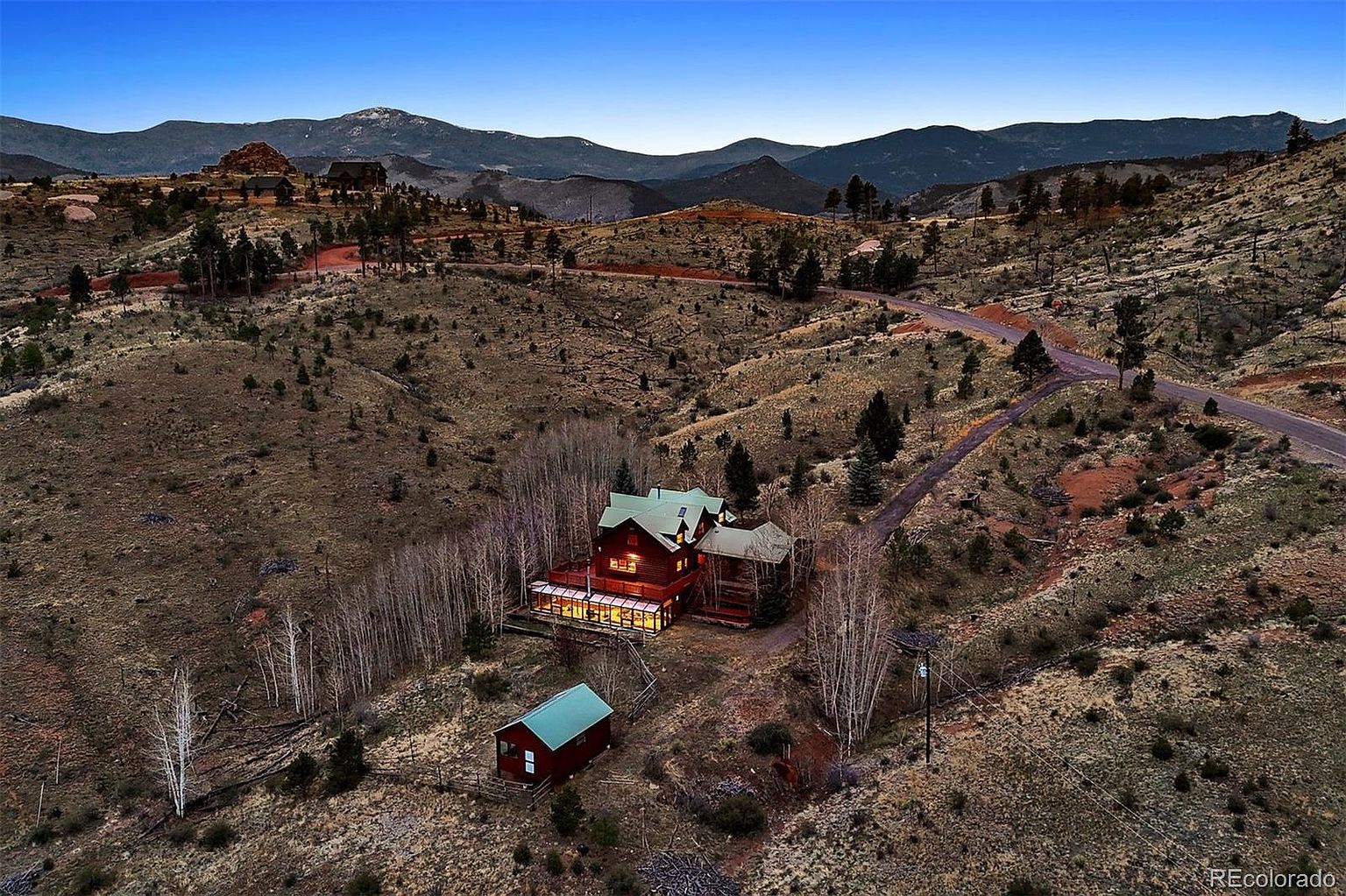 This aerial shot showcases a secluded mountain home with a rustic charm. The house features a green roof and a spacious deck, nestled among aspen trees and rolling hills. A separate small building is also visible, adding to the property's appeal and potential as a private retreat.
