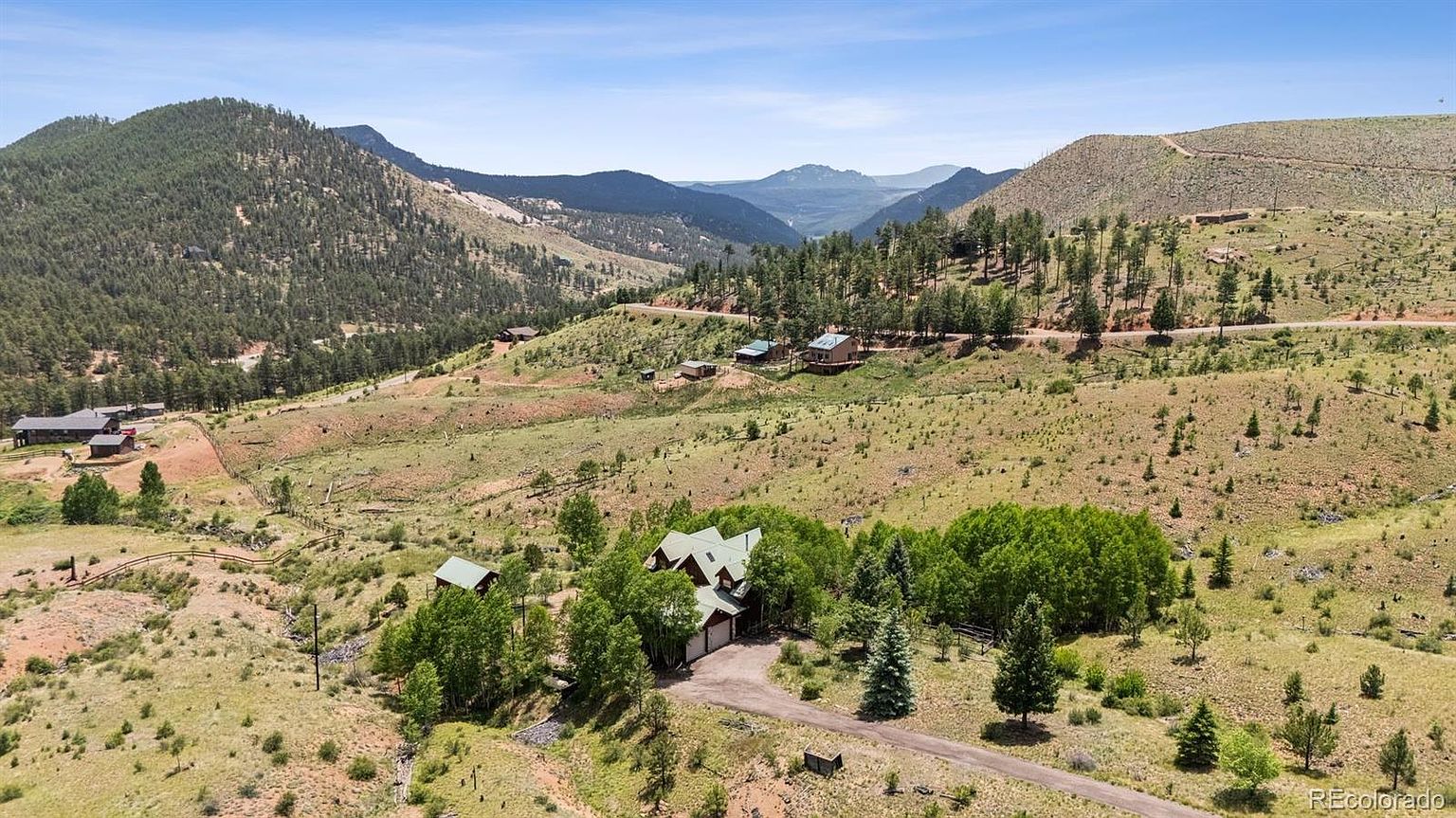 This aerial view showcases a beautiful property nestled in a mountainous landscape. The house, surrounded by lush greenery and mature trees, features a multi-car garage and a winding driveway. The overall impression is one of seclusion and natural beauty, ideal for a buyer seeking a private retreat.