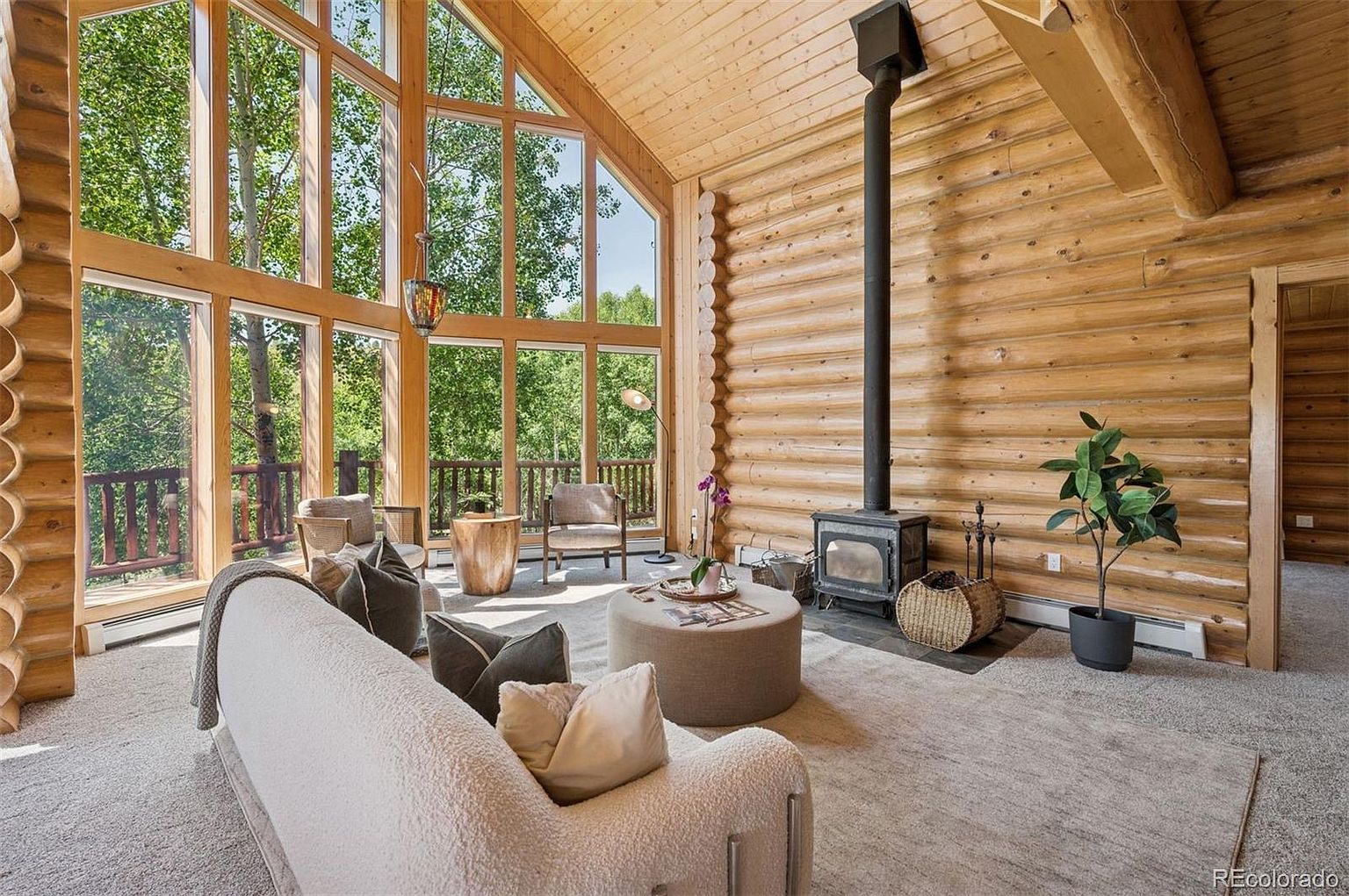 This is an interior shot of a cozy living room in a log cabin. The room features a large window wall offering views of the surrounding trees, a wood-burning stove with a black chimney, and comfortable seating including a white sofa and a round ottoman. The walls are made of logs, creating a warm and rustic atmosphere.