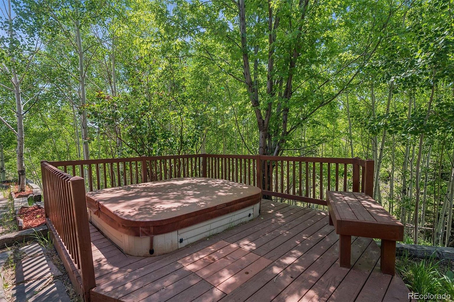 This image showcases a wooden deck featuring a covered hot tub and a bench, surrounded by lush greenery. The deck is enclosed by a wooden railing, providing a sense of privacy and seclusion. The scene evokes a relaxing and tranquil atmosphere, perfect for enjoying the outdoors.