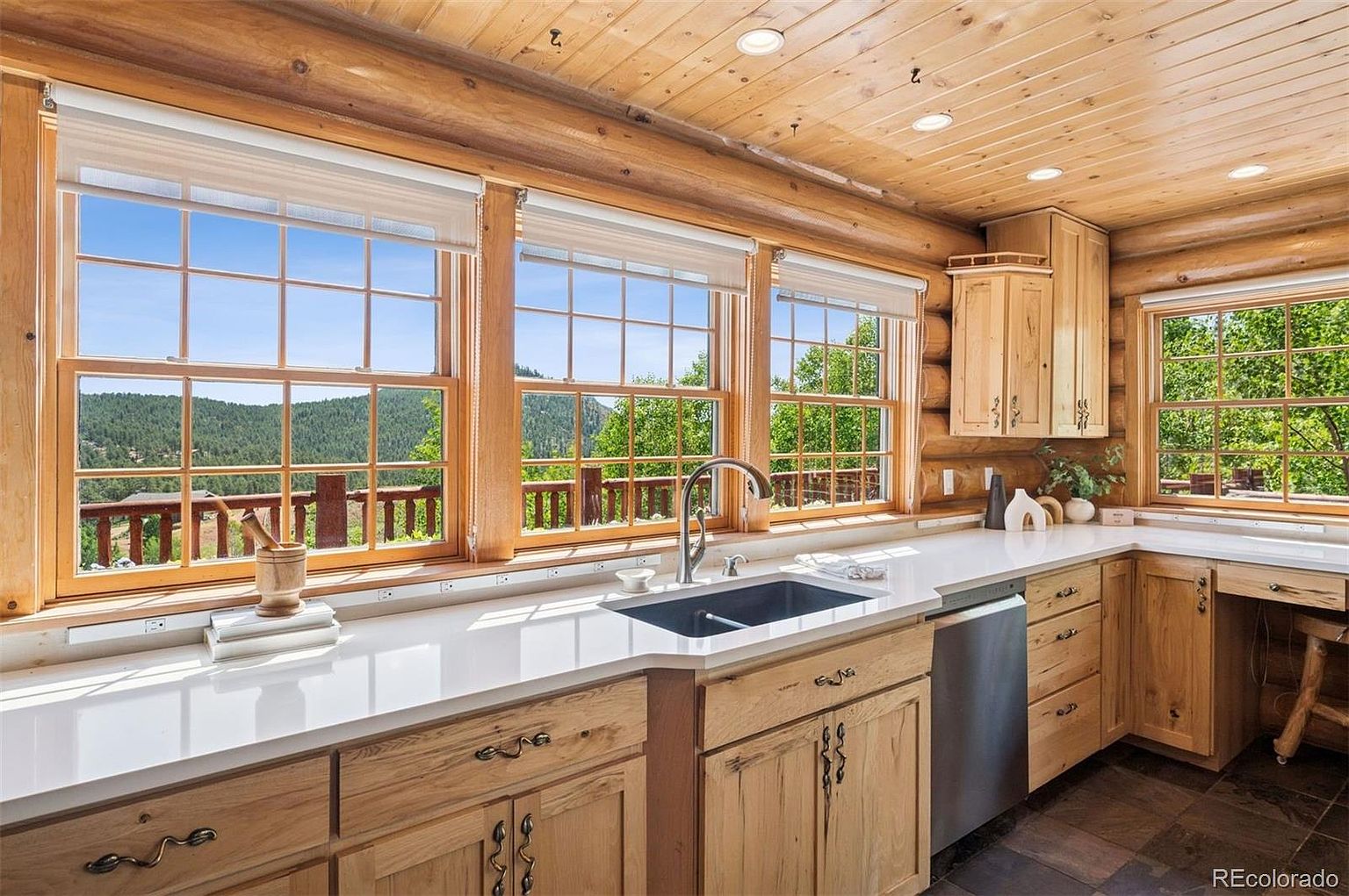This is a warm and inviting kitchen with a rustic log cabin aesthetic. The space features light wood cabinetry, a white countertop, and a dark sink. Large windows offer a scenic view of the outdoors, and the overall impression is cozy and well-lit.