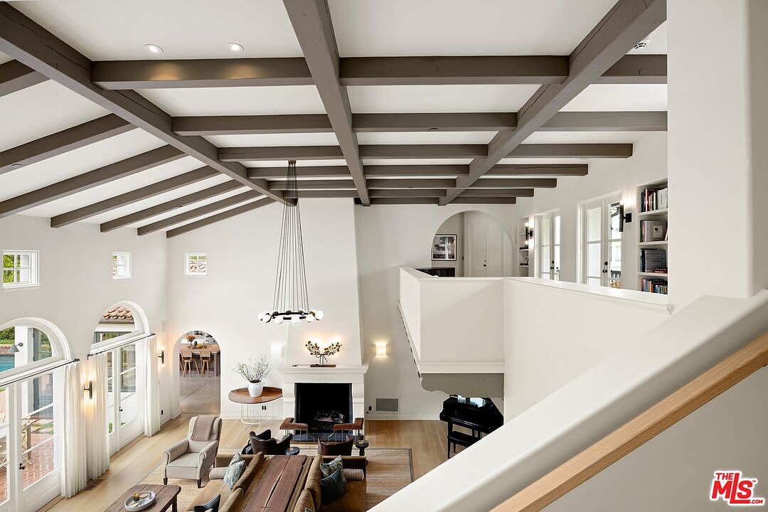 This is a high-angle shot of a grand living room featuring a high ceiling with exposed beams, a fireplace, and large windows that let in plenty of natural light. The room is furnished with a mix of modern and traditional pieces, including a large sofa, armchairs, and a piano on the side. The overall impression is one of spaciousness and elegance, perfect for entertaining or relaxing.
