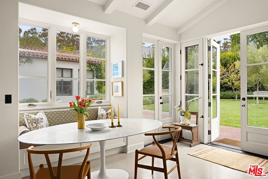 This is an interior shot of a dining room featuring an oval marble table with a white pedestal base, surrounded by wooden chairs with leather seats. A built-in bench with patterned cushions sits beneath a window, and French doors open to a lush green lawn, creating a bright and airy atmosphere. The room is painted in neutral tones, enhancing the natural light and highlighting the connection to the outdoors.