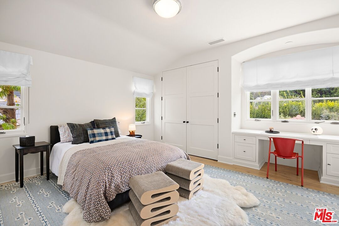 This is a bright and airy bedroom featuring a bed with patterned bedding, a side table, and a desk area with a red chair. The room has white walls, multiple windows with white shades, and a light blue patterned rug. The overall impression is clean, comfortable, and well-lit, making it an inviting space.