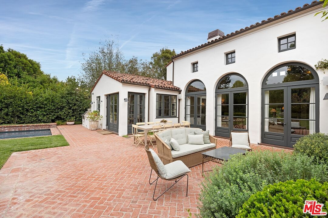 This image showcases a charming patio area of a home, featuring a herringbone brick pattern. The patio is furnished with a wicker sofa, chairs, and a coffee table, creating an inviting outdoor living space. The architecture includes arched windows and doors, complemented by lush greenery and a glimpse of a pool, enhancing the property's appeal.