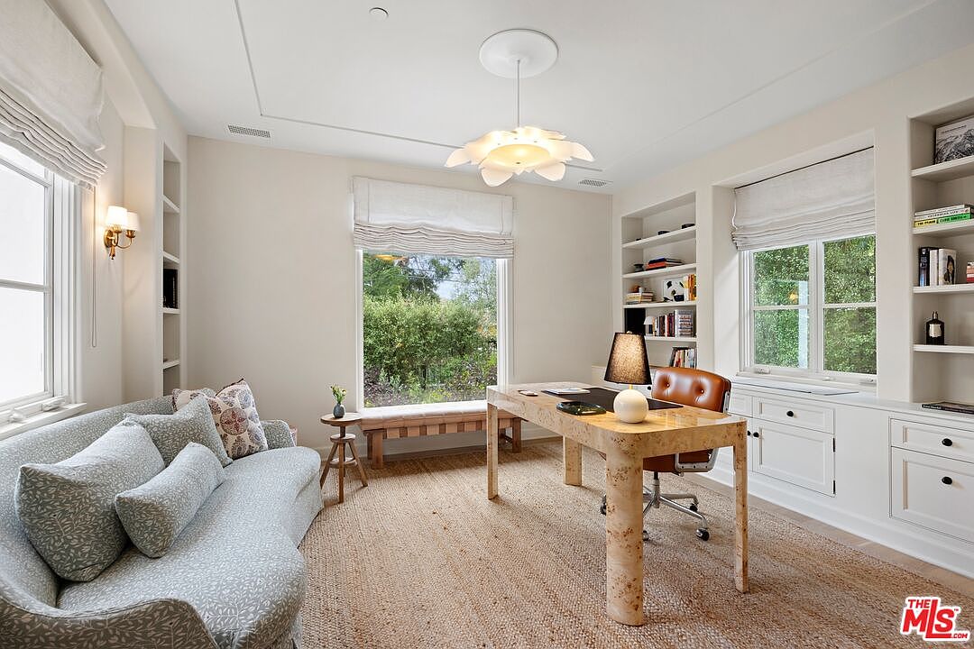 This is an interior shot of a well-lit home office. The room features built-in bookshelves, a large window with a view of greenery, and a unique desk with a leather chair. A comfortable sofa is placed near the window, creating a cozy and functional workspace.