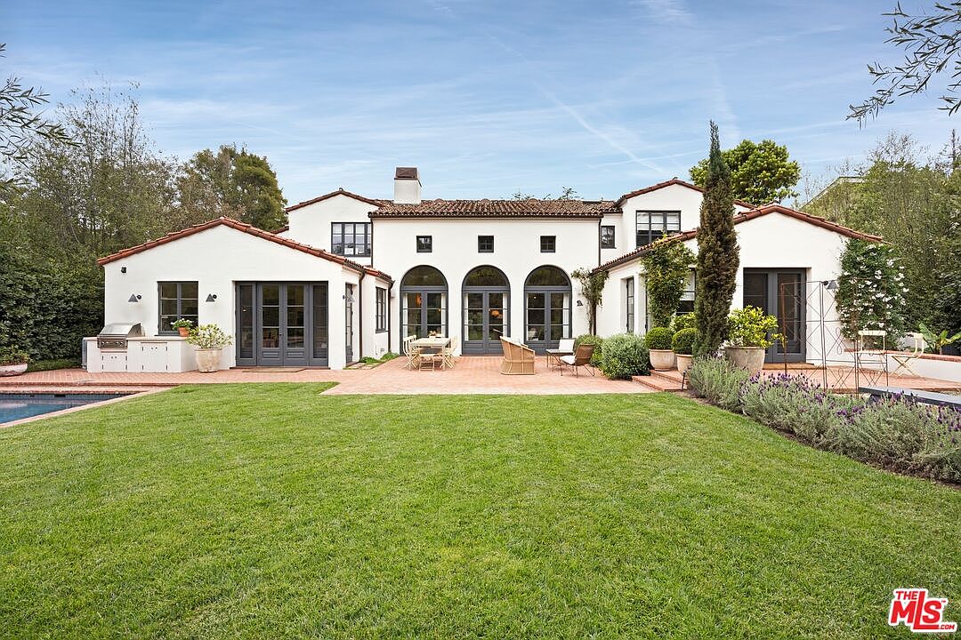 This image showcases the rear exterior of a Spanish-style home, featuring a white stucco facade, red tile roof, and dark-framed windows and doors. A well-manicured lawn extends from the house, leading to a brick patio area furnished with outdoor seating. The view emphasizes the home's architectural details and outdoor living space.
